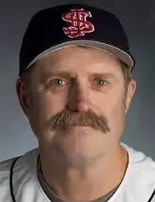 A middle-aged man with a mustache wears a dark baseball cap with red and white letters and a white baseball jersey, looking directly at the camera against a neutral background.